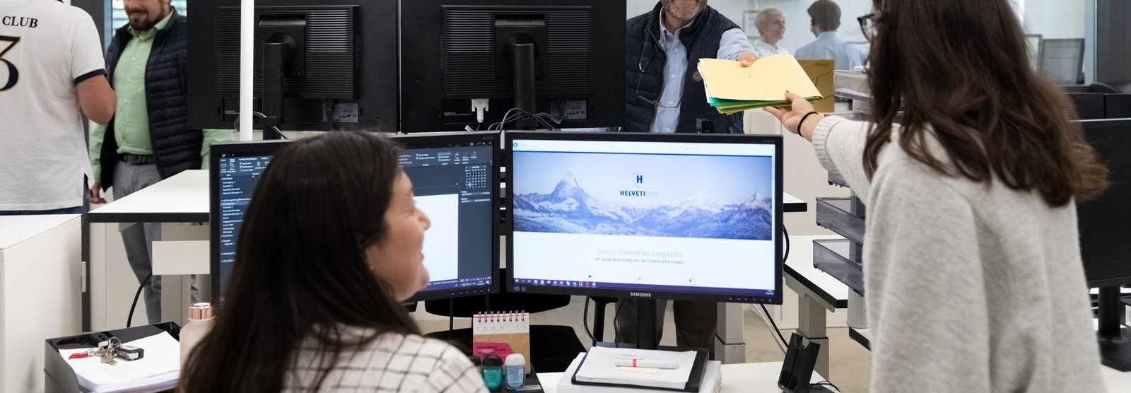 Mitarbeitende im Helveticor-Büro beim Austausch von Dokumenten; im Vordergrund mehrere Bildschirme, im Hintergrund freundliche Interaktion