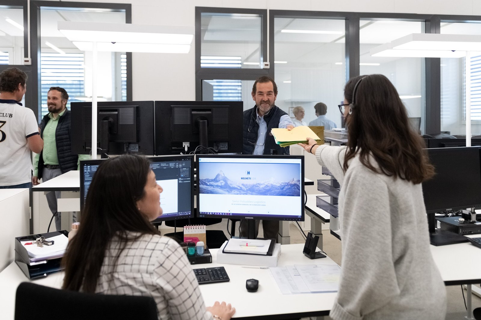 Mitarbeitende im Helveticor-Büro beim Austausch von Dokumenten; im Vordergrund mehrere Bildschirme, im Hintergrund freundliche Interaktion