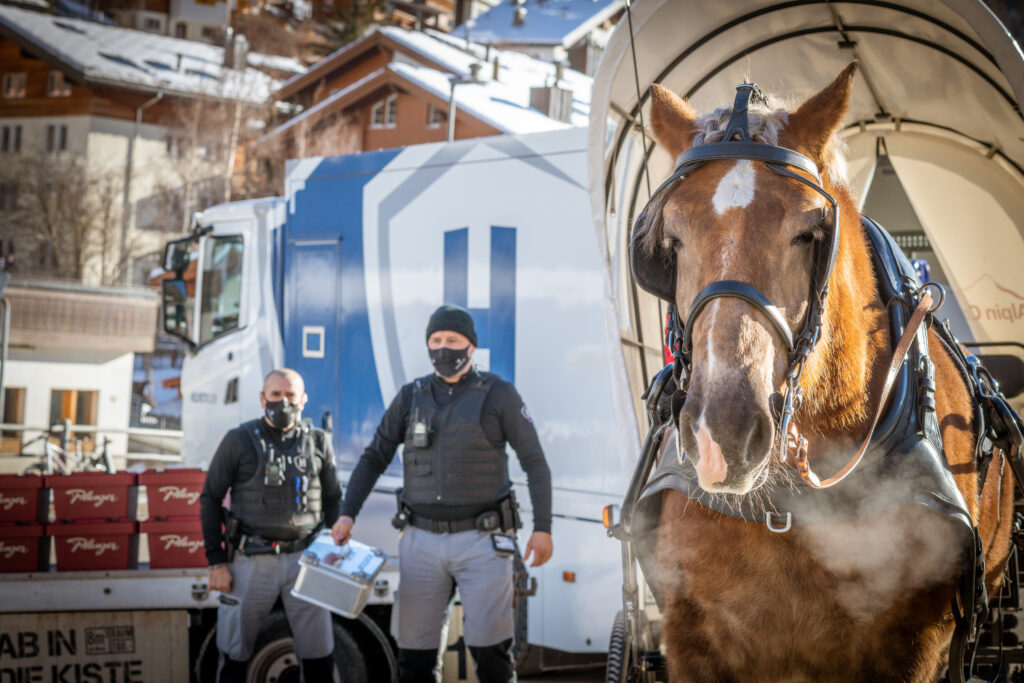 Zwei Helveticor-Mitarbeiter mit Sicherheitsausrüstung stehen neben einem gepanzerten Transportfahrzeug, im Vordergrund eine dampfende Pferdekutsche – Logistik in Zermatt